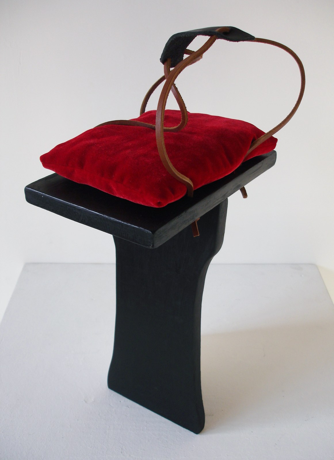 La sculpture : un socle de bois noir laqué à colonne galbée porte un coussin de velours rouge ; au-dessus, des lanières de cuir fauve s'élèvent en boucles et se rejoignent sous un arceau, dessinant une cage ouverte.