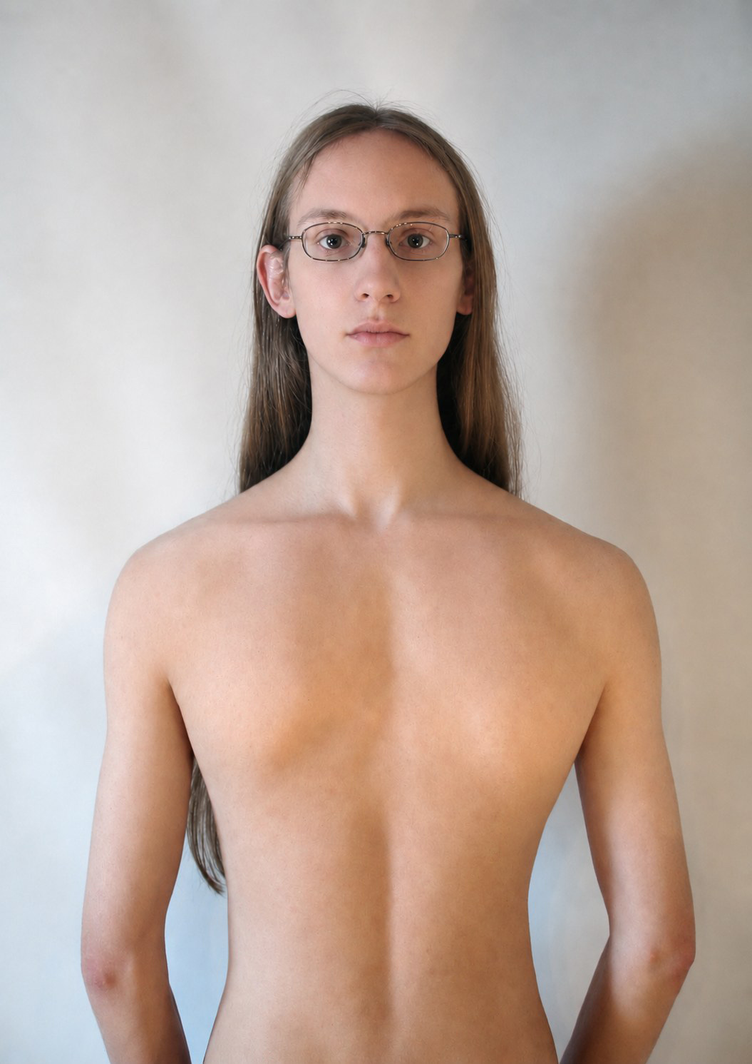 Autoportrait en buste sur fond clair&nbsp;: une personne aux cheveux longs et aux lunettes fines regarde l'objectif, torse nu. Sous les clavicules, ce n'est pas la poitrine que l'on voit mais le dos — un creux vertical le long de la colonne, sans aucune couture apparente.