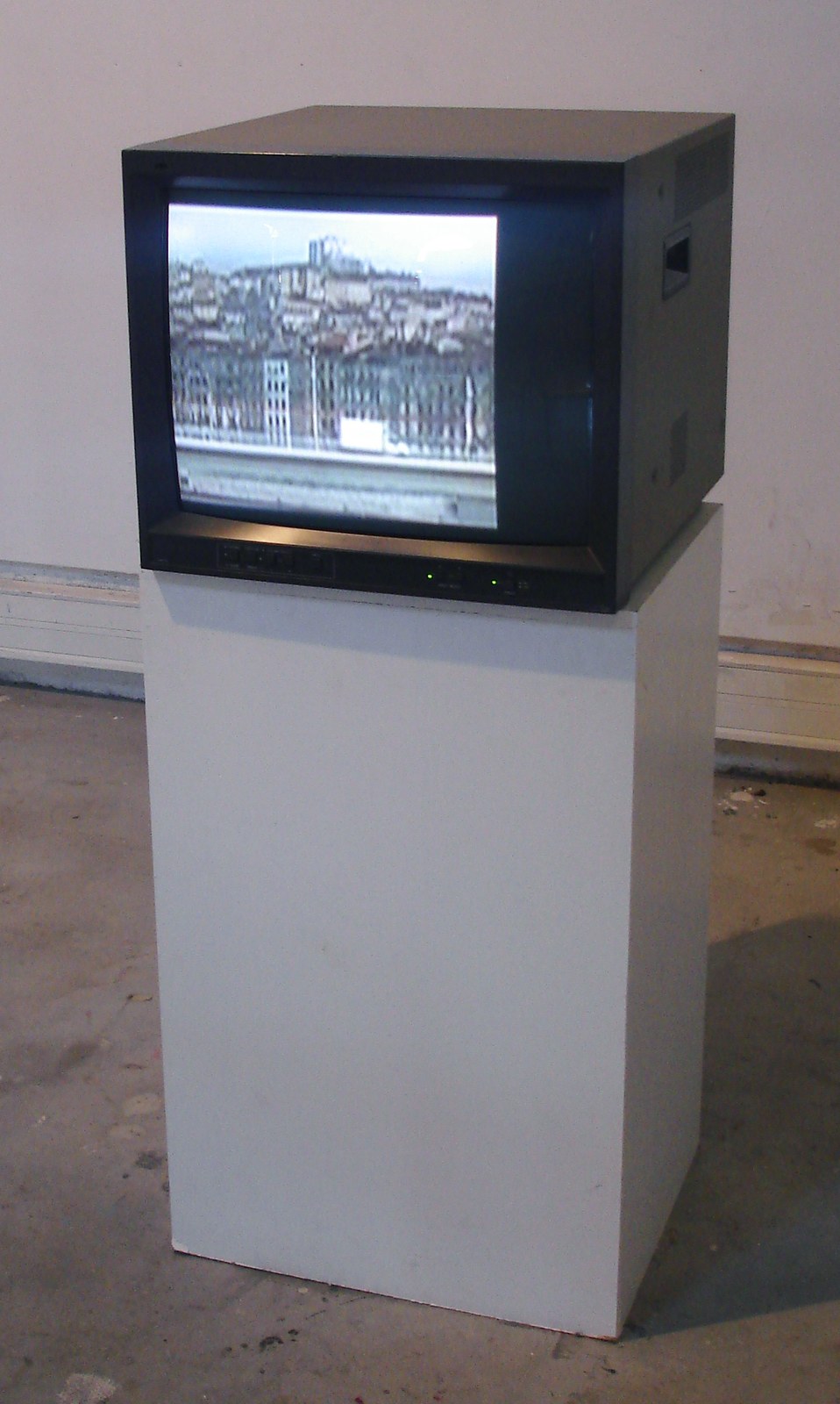 Vue d'installation&nbsp;: un téléviseur cathodique noir, au format 4/3, posé sur un socle blanc à hauteur de poitrine, dans une salle au sol de béton et au mur clair. L'écran allumé montre une image grise et tremblante — une ville étagée à flanc de colline, ses façades serrées au-dessus d'un quai.