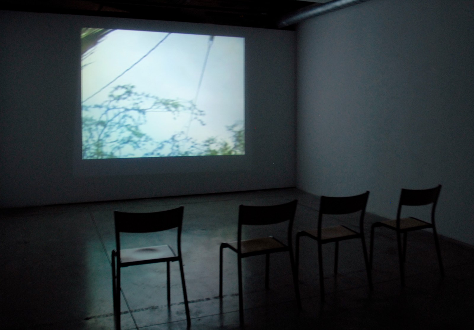 Vue d'installation dans une salle obscure au sol de béton&nbsp;: sur le mur du fond, une projection lumineuse montre un ciel pâle traversé de câbles et de branches, l'image d'un espace recomposé. Quatre chaises d'écolier en bois et métal sont alignées devant, vides, tournées vers le mur.
