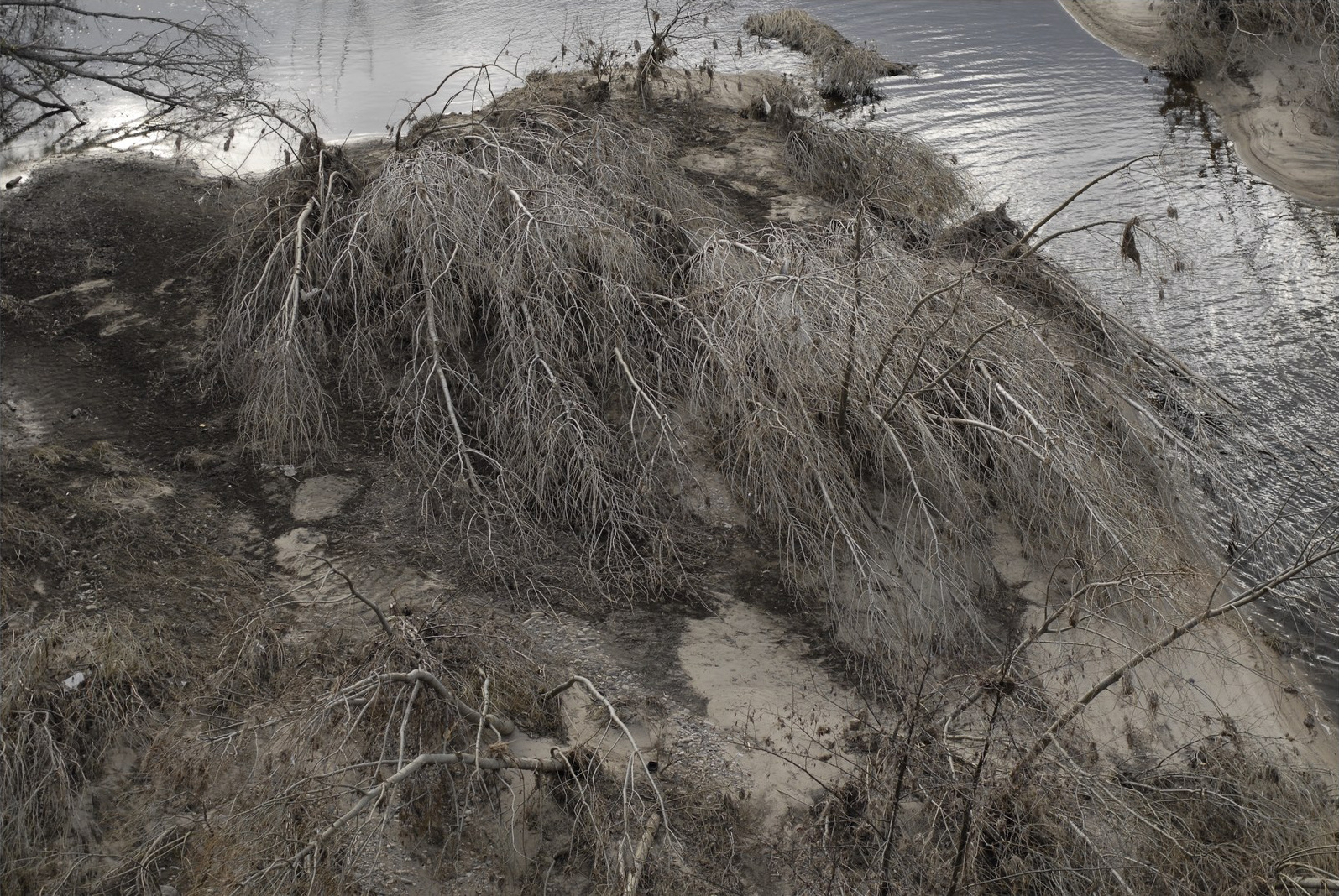 Tirage du bas&nbsp;: vue plongeante, presque à la verticale, sur un talus entièrement peigné par la crue — un enchevêtrement d'arbres couchés et de branches sèches recouvrant le sable, l'eau ridée occupant le haut du cadre.