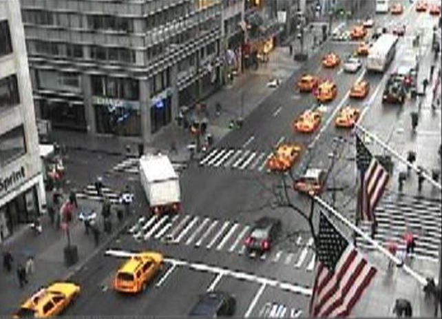 Seconde séquence, capture d'une webcam publique de New York&nbsp;: vue en plongée d'un carrefour de Manhattan un jour de pluie, l'asphalte luisant strié de passages piétons blancs. Une douzaine de taxis jaunes remontent l'avenue, un camion de livraison blanc attend au feu&nbsp;; au premier plan à droite, deux drapeaux américains pendent le long de leurs hampes.
