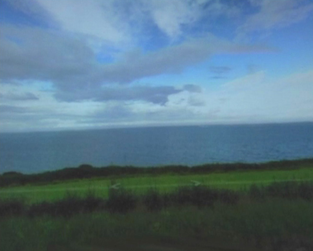 Photogramme du même défilement, la vue dégagée&nbsp;: un ciel de nuages bas occupe la moitié haute, la mer en dessous forme une lame pâle et plate, puis une bande d'herbe vert vif et le talus sombre du bord de route. De fines lignes horizontales blanches traversent l'image, trace du balayage vidéo.