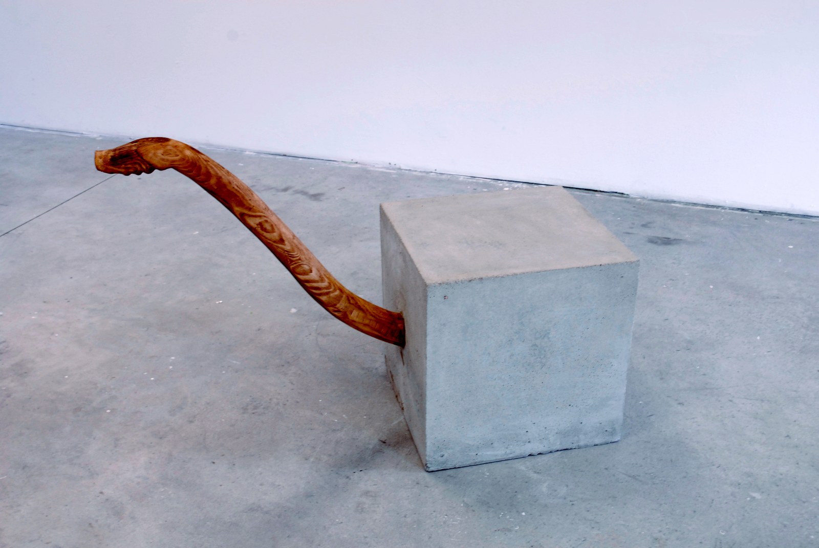 Vue d'ensemble au sol&nbsp;: un cube de béton gris clair d'une cinquantaine de centimètres, d'où sort à mi-hauteur un long manche de bois clair et veiné, courbe comme un bras, dont l'extrémité s'épaissit puis s'arrondit. L'ensemble repose sur une dalle de béton, devant un mur blanc.