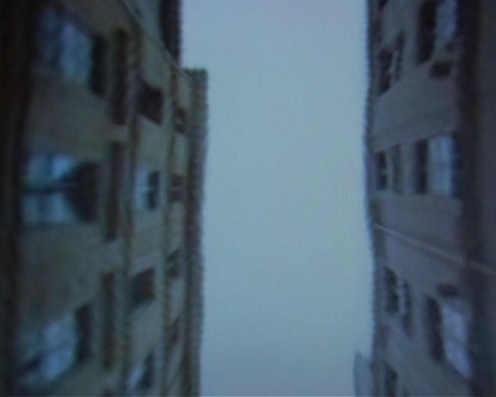 Contre-plongée entre deux immeubles, filmée à même l'écran&nbsp;: les façades penchent de part et d'autre du cadre, floues et bleutées, et laissent au centre une bande de ciel blanc, verticale, sans nuage. Les fenêtres ne sont plus que des rectangles sombres étirés.