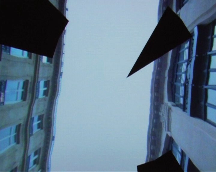 Contre-plongée nette entre deux immeubles haussmanniens&nbsp;: les façades de pierre claire, leurs volets et leurs ferronneries, s'inclinent de chaque côté d'une bande de ciel blanc. De grands triangles noirs, aux bords parfaitement rectilignes, mordent l'image en haut à gauche, à droite et en bas — les zones que la machine n'a pas photographiées.