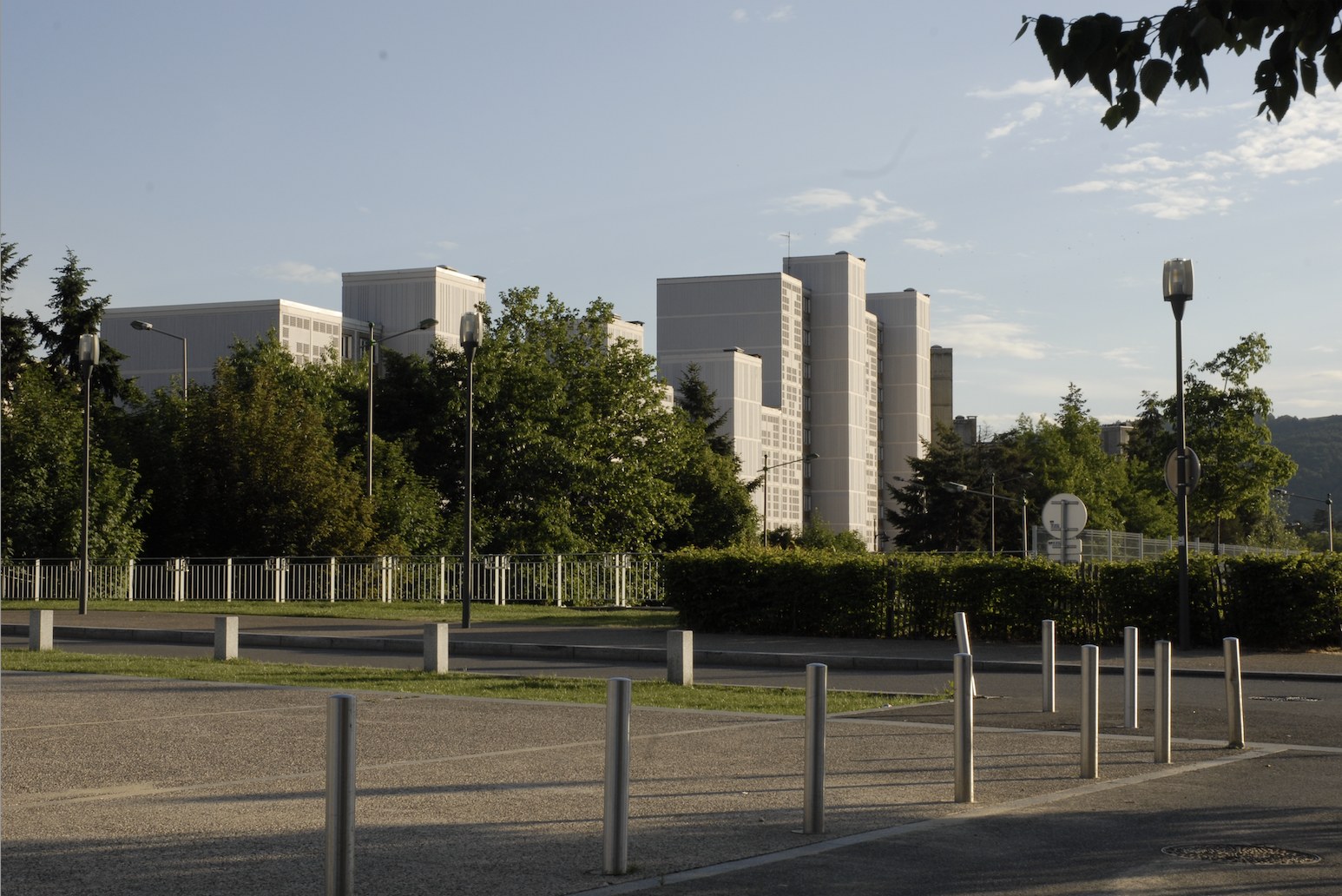En fin de journée, un ensemble de barres et de tours de logements aux façades blanches se détache au-dessus d'une masse d'arbres qui en masque le pied. Au premier plan, une esplanade de béton bordée de bornes et de potelets métalliques, une bande d'herbe, une haie taillée et des lampadaires&nbsp;; au loin, à droite, la ligne des collines.