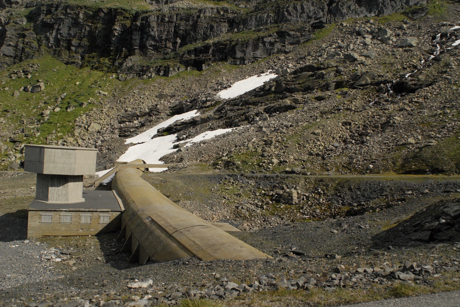Dans un versant de montagne fait d'éboulis, de roche noire et de plaques de neige, une conduite forcée en béton descend en diagonale et vient buter sur une petite centrale&nbsp;: un bâtiment bas à fenêtres carrées, surmonté d'un bloc de béton en encorbellement. Un filet d'eau dévale la pente à droite&nbsp;; une piste de gravier traverse le bas du cadre.
