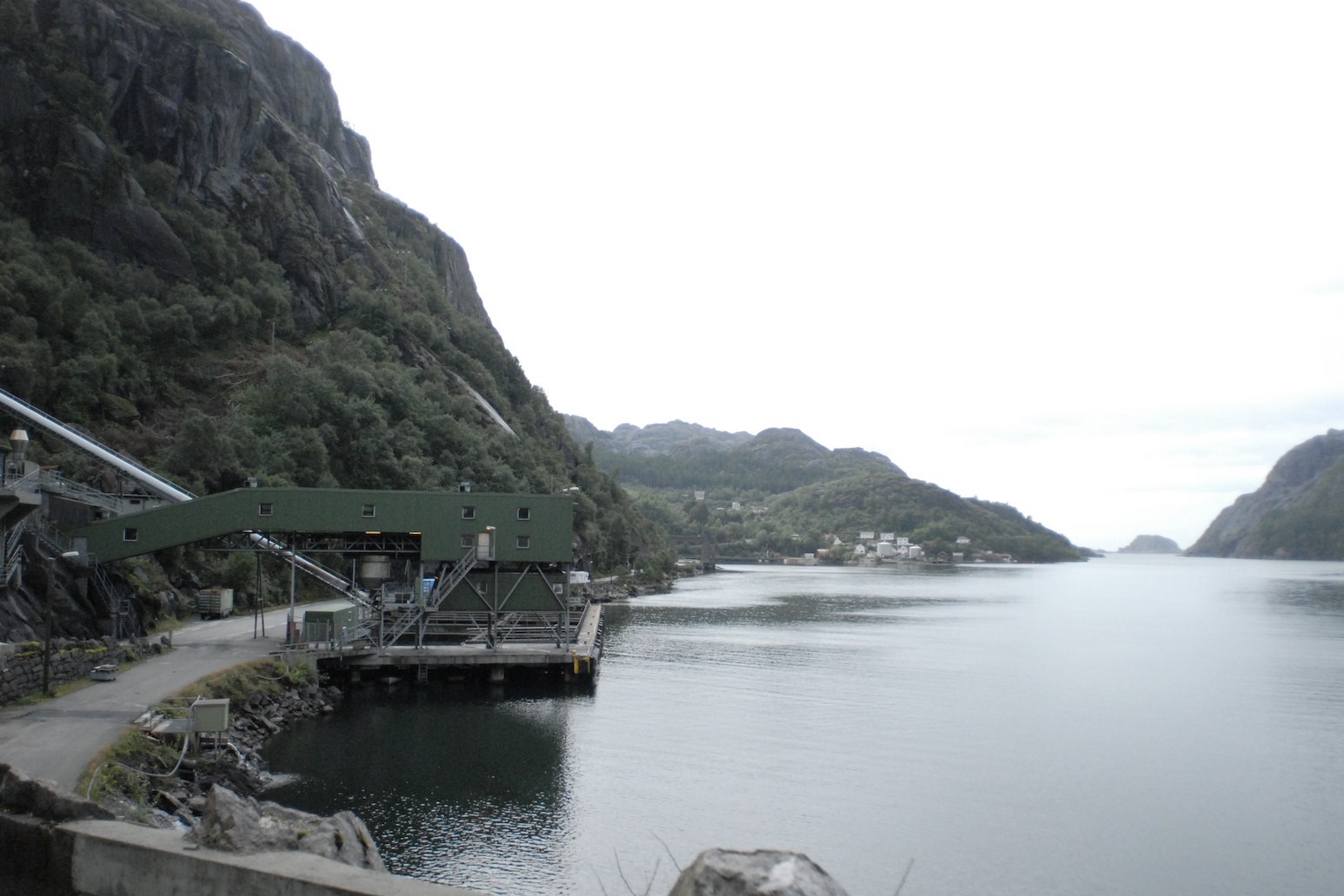 Au bord d'un fjord aux eaux plates et grises, une installation industrielle vert sombre est posée sur pilotis contre une falaise boisée&nbsp;: un long tapis convoyeur descend de la roche jusqu'au bâtiment, qui avance sur un quai. À l'arrière-plan, la rive opposée, quelques maisons blanches au pied des versants, et le ciel blanc de la brume.