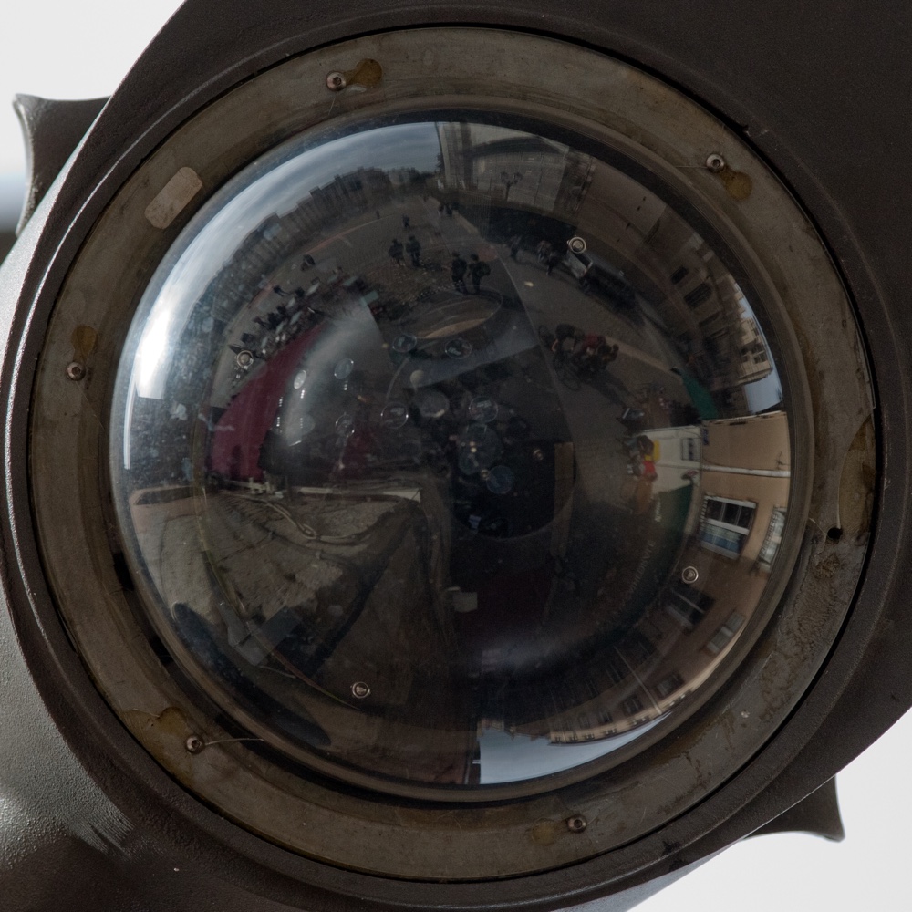 Dôme de verre d'une caméra de vidéosurveillance au boîtier gris-brun bagué d'acier et constellé de vis&nbsp;: le verre bombé réfléchit la place en anneau — façades pliées vers le pourtour, une terrasse rouge, des passants minuscules, le ciel pâle au centre.
