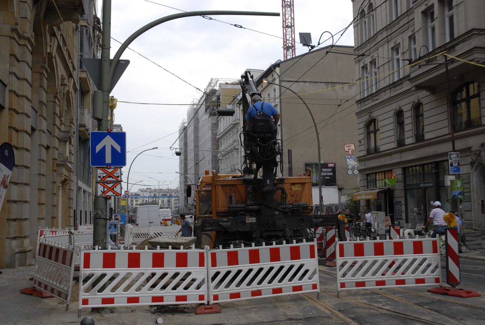 Une rue barrée par des barrières de chantier rouge et blanc. Au centre, une pelleteuse orange dont le bras hydraulique est dressé&nbsp;: un ouvrier en tee-shirt bleu se tient debout sur la machine, de dos, penché vers le mécanisme. Derrière, les caténaires du tramway, une grue rouge et blanche, des façades berlinoises&nbsp;; sur la chaussée éventrée, un bloc de béton et les rails à nu.