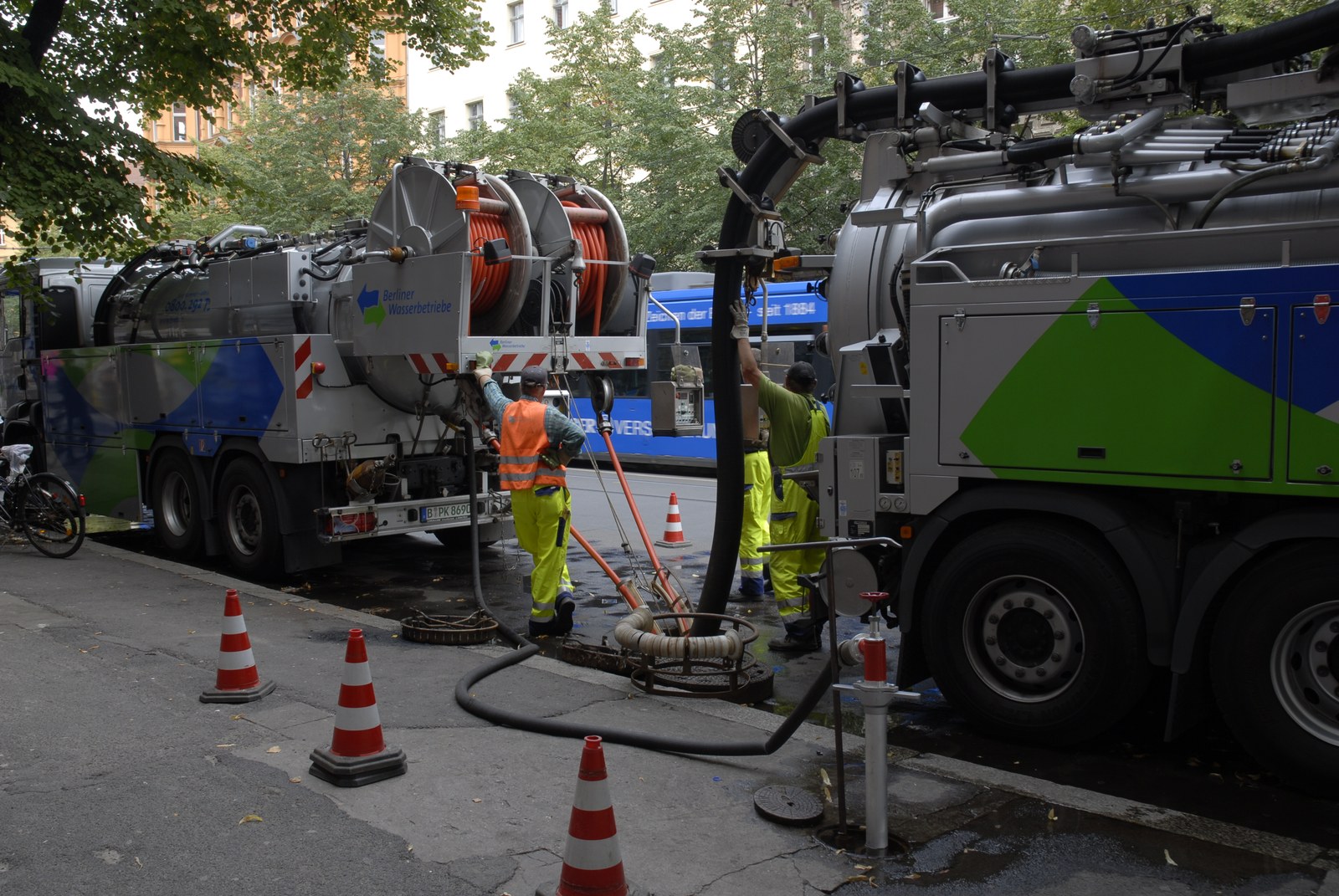 Deux camions hydrocureurs gris et verts des services des eaux, garés de part et d'autre d'une chaussée mouillée, reliés par de larges tuyaux à une bouche d'égout ouverte au centre du cadre. Deux ouvriers en pantalon jaune fluorescent manœuvrent les flexibles, l'un de dos, l'autre bras levé. Des cônes de signalisation blancs et rouges bordent le trottoir&nbsp;; un tramway bleu passe à l'arrière-plan.