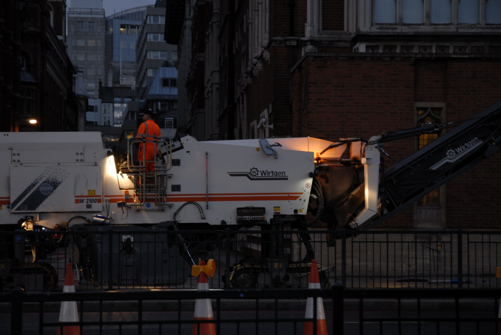 À la nuit tombante, une raboteuse de chaussée blanche et orange est arrêtée derrière une barrière métallique, ses phares allumés. Debout sur la passerelle de la machine, un ouvrier en combinaison orange fluorescent regarde vers l'avant, de dos. Autour, les façades sombres d'un quartier d'affaires, une fenêtre encore éclairée&nbsp;; au premier plan, des cônes de signalisation.