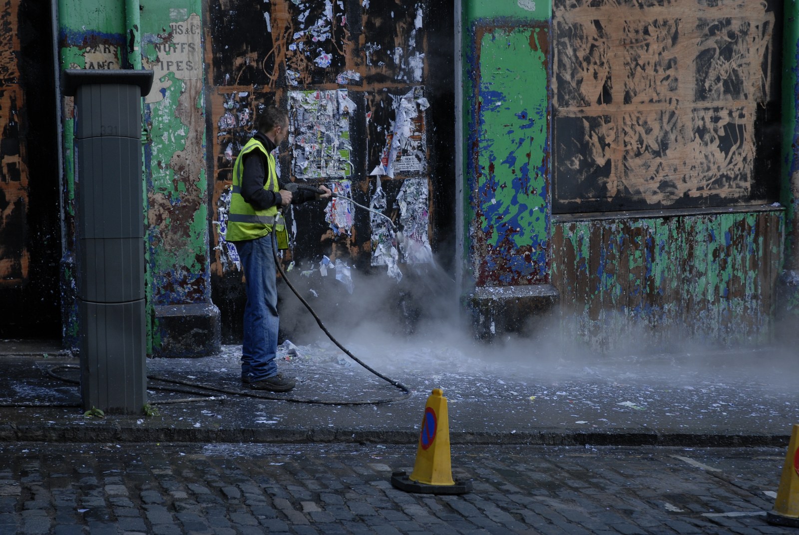 Devant une devanture condamnée, couverte de couches d'affiches lacérées et de peinture écaillée verte, bleue et noire, un homme en gilet jaune fluorescent dirige une lance à haute pression vers le mur. Le jet lève un nuage de vapeur et projette des lambeaux de papier au sol, sur les pavés mouillés. À droite, un cône de signalisation jaune.