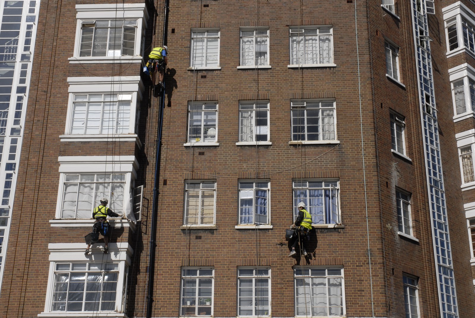 Façade de brique d'un immeuble de logements, vue en contre-plongée. Trois cordistes en gilet fluorescent y sont suspendus à des cordes, casque blanc, seau accroché au harnais, chacun à une hauteur différente&nbsp;: deux devant les fenêtres à meneaux blancs, un troisième contre la descente d'eaux pluviales. Derrière les vitres, des rideaux, une plante, une vie ordinaire.