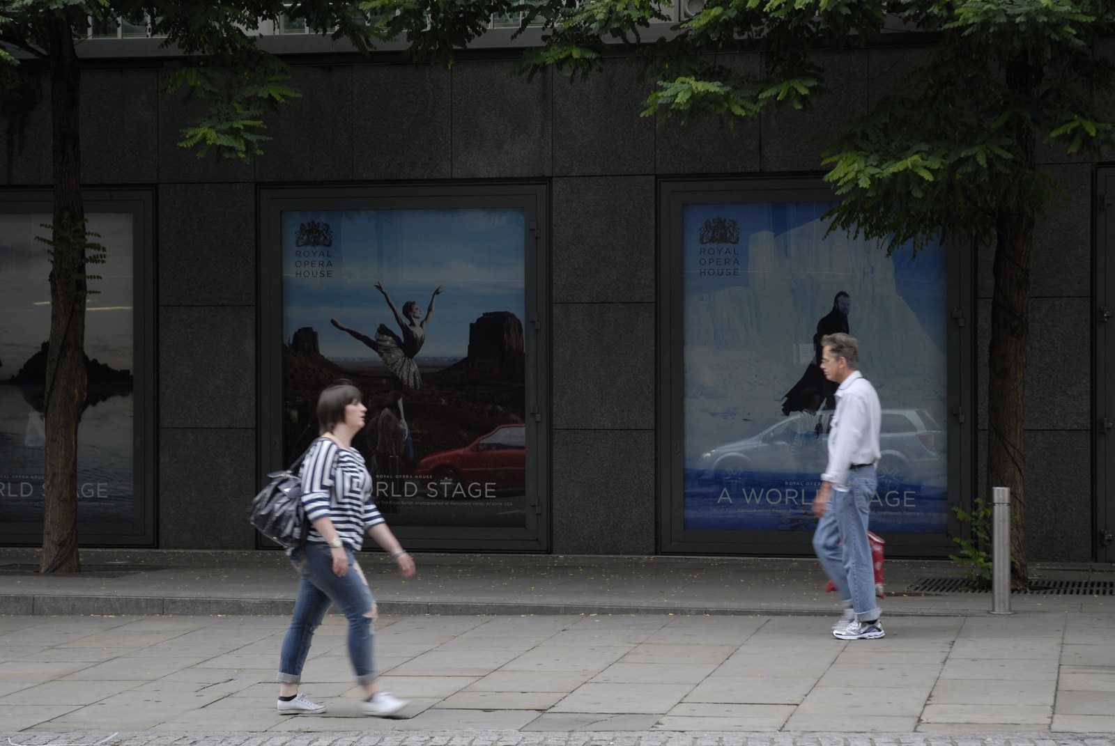 Devant une façade de granit noir, deux affiches rétroéclairées du Royal Opera House&nbsp;: sur l'une, une danseuse en tutu bondit bras levés au milieu d'un désert de buttes rouges&nbsp;; sur l'autre, une silhouette sombre se tient au bord d'un rivage de glace. Au bas de chacune, la ligne «&nbsp;A WORLD STAGE&nbsp;». Une femme en marinière, sac de voyage à l'épaule, traverse le trottoir sans regarder les affiches.