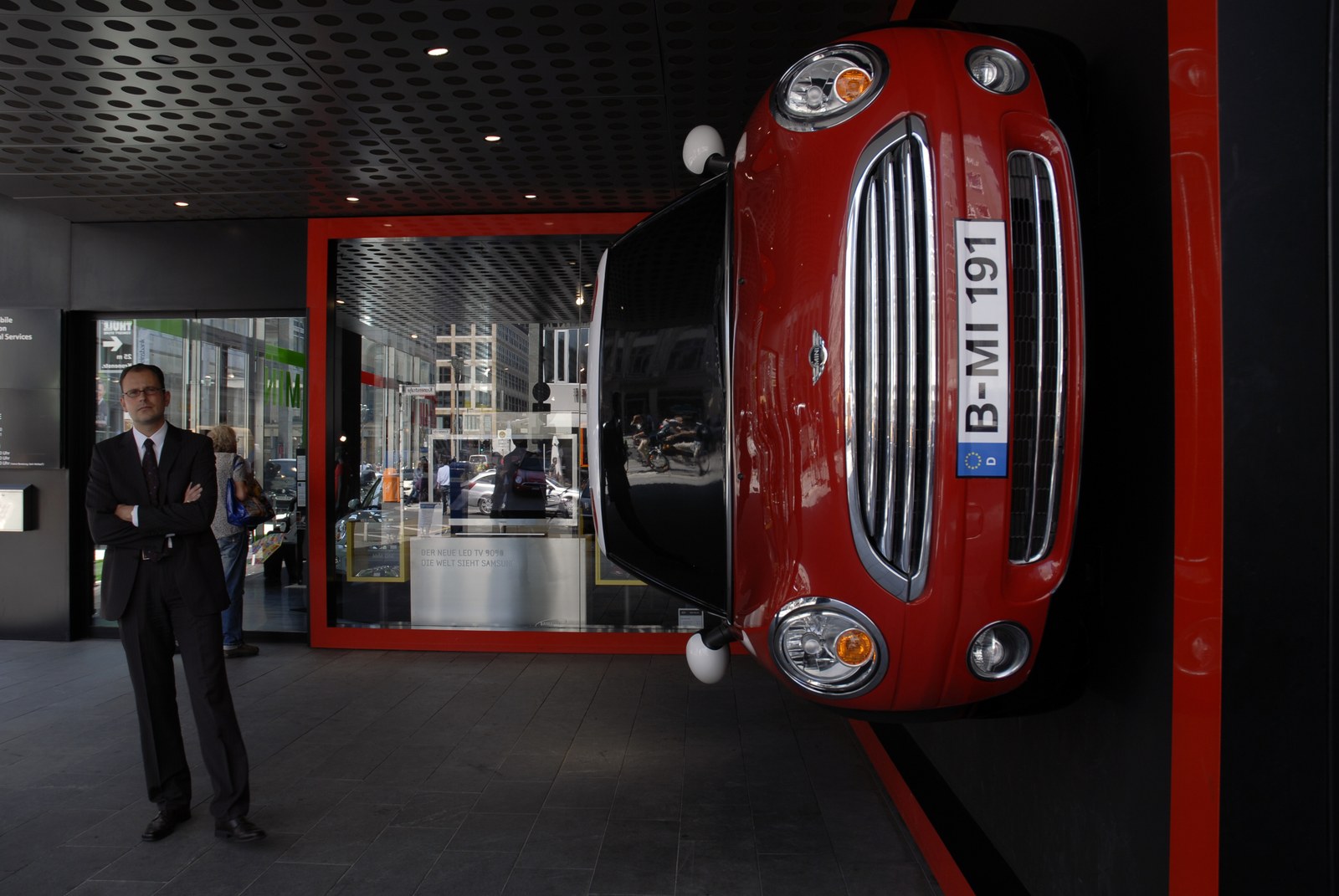 Sous l'auvent noir d'un hall vitré, une petite voiture rouge est fixée à la verticale contre le mur, roues dans le vide, calandre chromée en avant et plaque d'immatriculation berlinoise à hauteur des yeux. À gauche, devant la vitrine, un homme en costume sombre attend, bras croisés, tourné vers la rue&nbsp;; il ne la regarde pas.