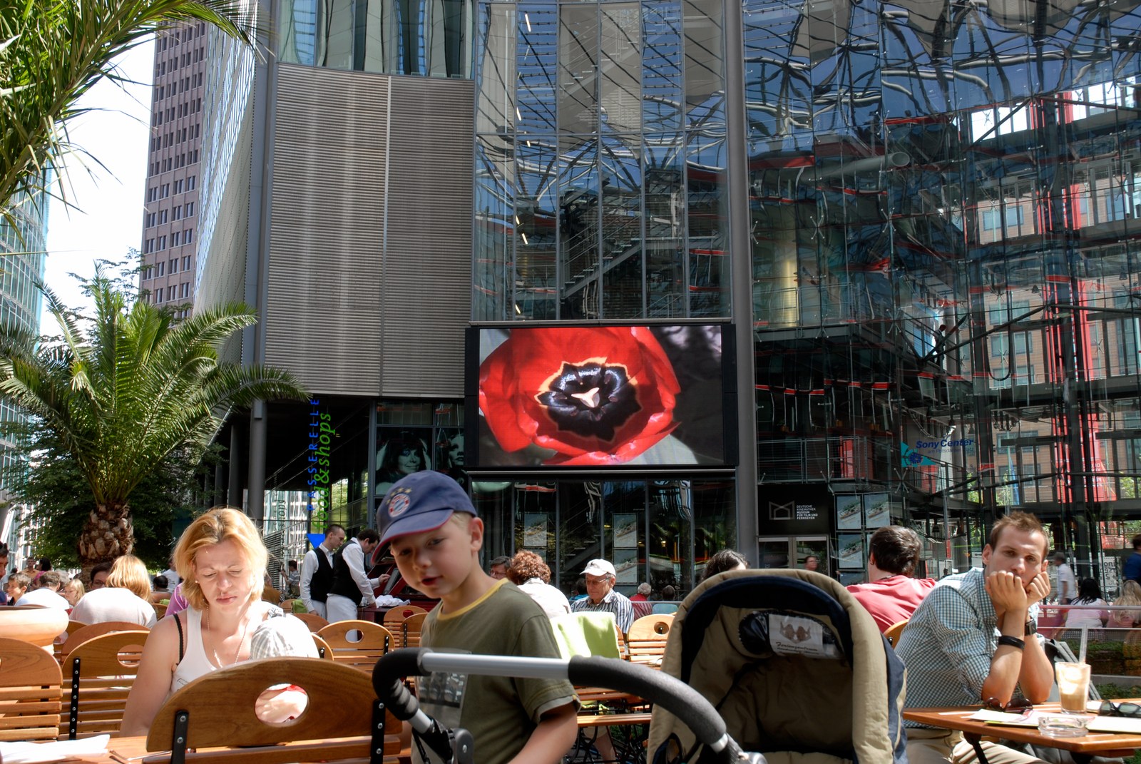 Terrasse de café bondée sous les verrières d'un centre commercial berlinois. Au-dessus des tables, un grand écran allumé montre en gros plan une tulipe rouge ouverte, cœur noir. Aux tables, une femme baisse les yeux, un enfant en casquette pousse une poussette, un homme accoudé regarde ailleurs.