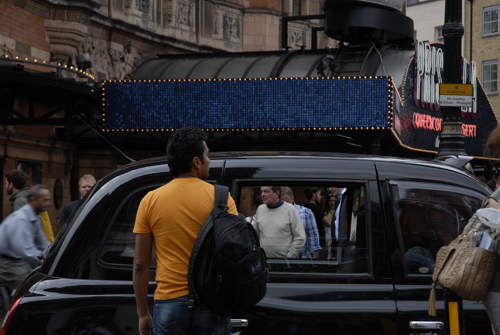 Rue de Londres devant un théâtre&nbsp;: un taxi noir occupe l'avant-plan, l'enseigne lumineuse du spectacle et un bandeau d'ampoules au-dessus. Un homme en tee-shirt orange, sac à dos, passe de dos&nbsp;; dans la vitre du taxi se reflètent les visages et les corps des passants du trottoir opposé.