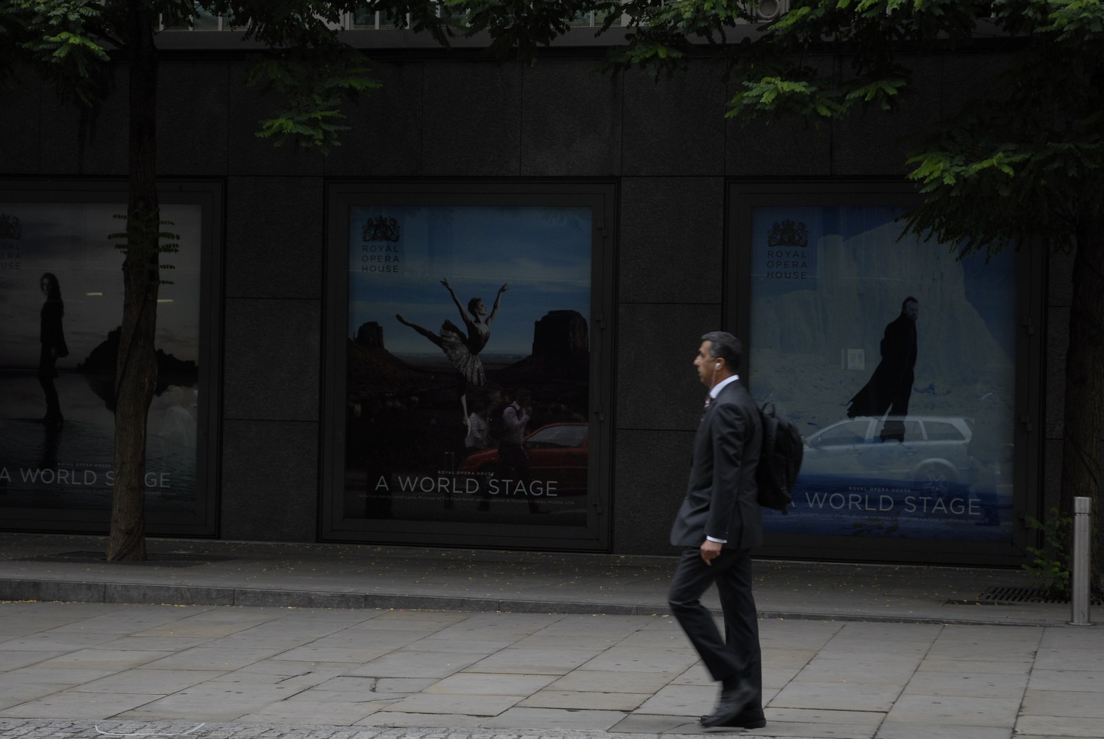 La même façade de granit noir et ses affiches «&nbsp;A WORLD STAGE&nbsp;»&nbsp;: la danseuse du désert au centre, la silhouette du rivage de glace à droite. Un homme en costume sombre, écouteur à l'oreille et sac à l'épaule, traverse le cadre à hauteur des danseurs, le regard droit devant lui.