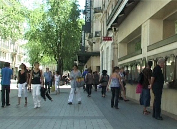 Photogramme de la déambulation&nbsp;: une rue commerçante de Lyon un jour d'affluence, filmée à hauteur d'épaule. Sur la droite, la façade de pierre claire d'un grand magasin et ses vitrines&nbsp;; à gauche, une rangée d'arbres. Une vingtaine de passants avancent en tous sens sur le trottoir dallé, sacs à la main&nbsp;; au centre, une femme en tunique claire marche vers la caméra.