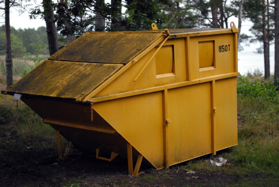 Benne à ordures jaune, capot bâché relevé et deux trappes carrées, marquée « 850T », posée dans un sous-bois de pins au bord d'une étendue d'eau.