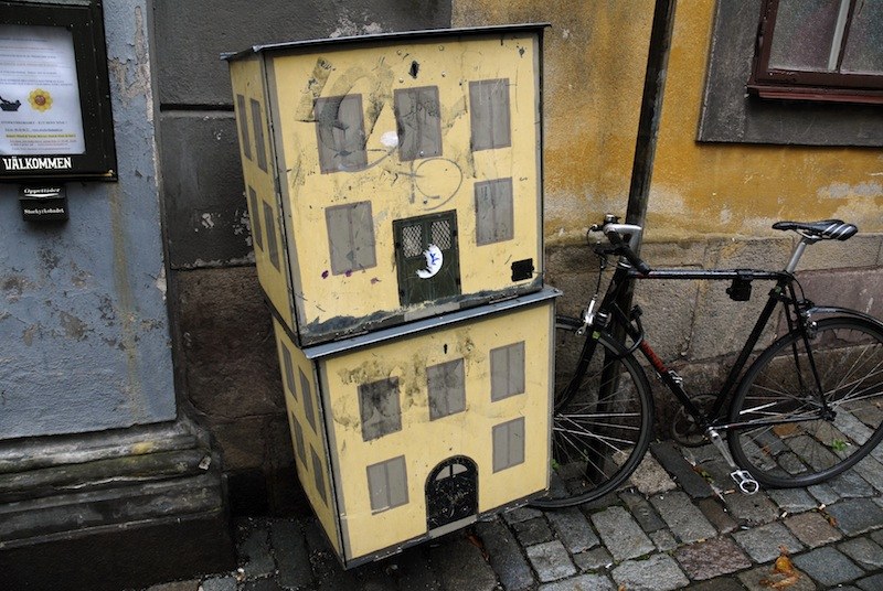 Coffret technique peint en maison jaune à deux étages, fenêtres et porte figurées au pochoir, adossé au mur d'une ruelle pavée, un vélo appuyé contre lui.