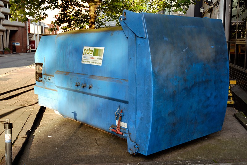 Compacteur à déchets bleu, capot bombé et flancs marqués d'une étiquette, posé sur le trottoir d'une rue commerçante, à hauteur d'homme.