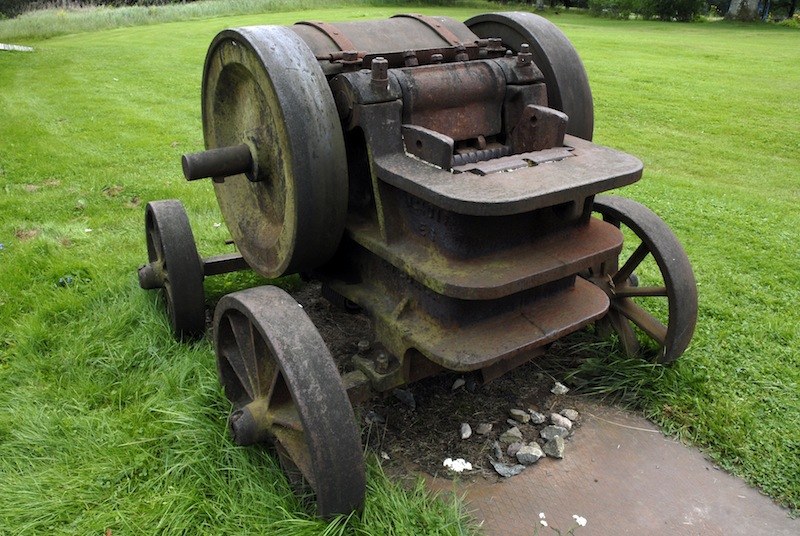 Vieille machine agricole en fonte rouillée, deux grands volants et deux roues à rayons, ses gradins de métal empilés comme des marches, sur une pelouse tondue.