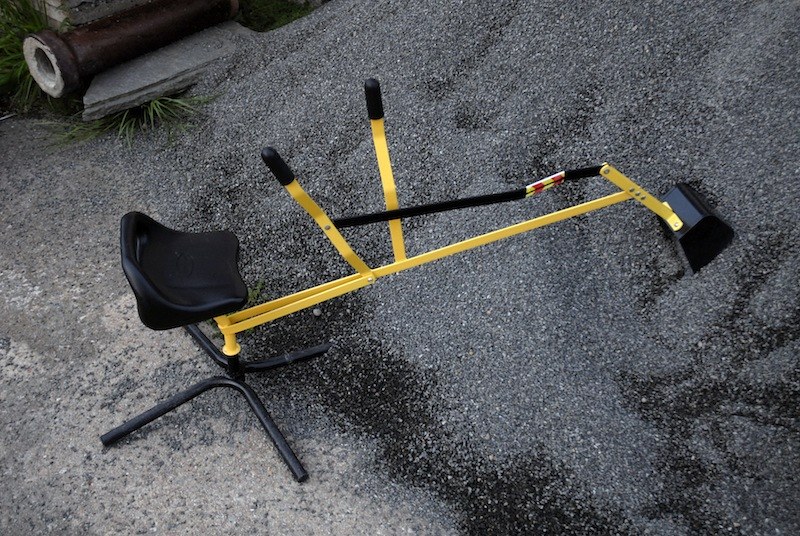 Structure tubulaire jaune à selle noire montée sur pied, longue flèche articulée terminée par un godet&nbsp;: un jeu de bac à sable, couché sur un tas de gravier.