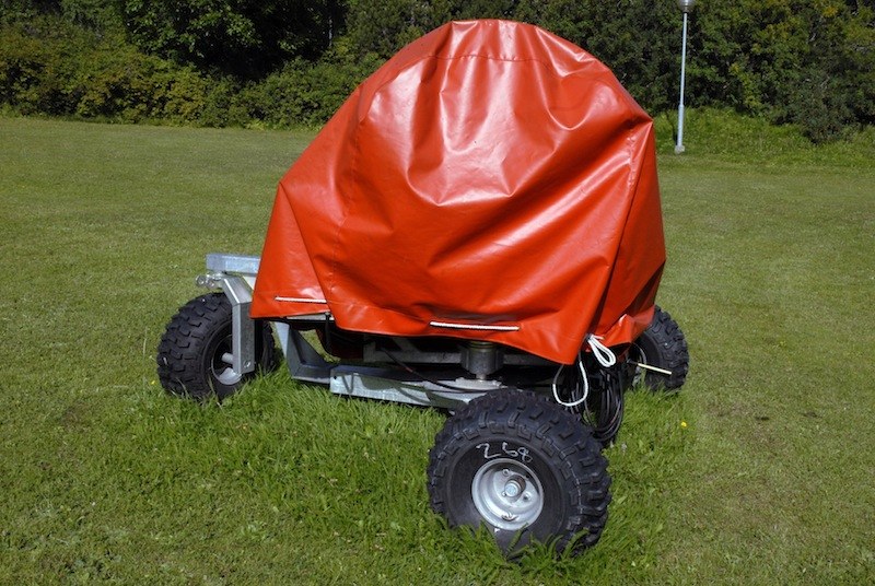 Remorque à quatre grosses roues à crampons, sa charge entièrement enveloppée d'une bâche rouge vif, immobile au milieu d'une pelouse.