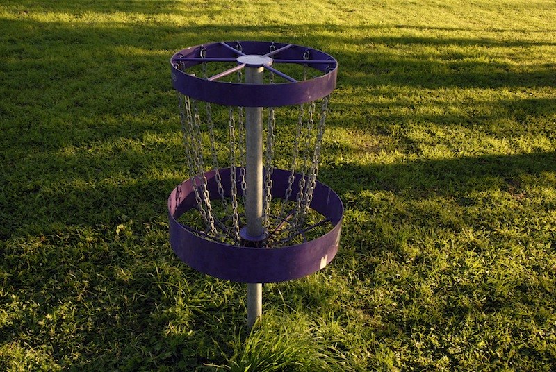 Corbeille de disc-golf violette&nbsp;: deux couronnes de tôle superposées, reliées par un rideau de chaînes, sur un mât planté dans une pelouse en lumière rasante.