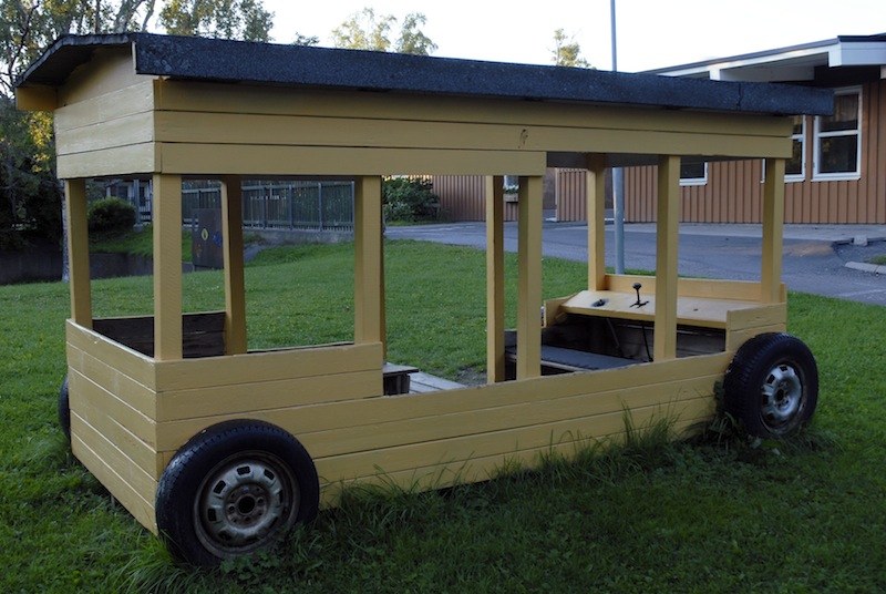 Grande structure de jeu en bois peint en jaune, figurant un autobus&nbsp;: toit noir en débord, montants, banquettes et volant, montée sur quatre roues de voiture, dans une cour d'école.