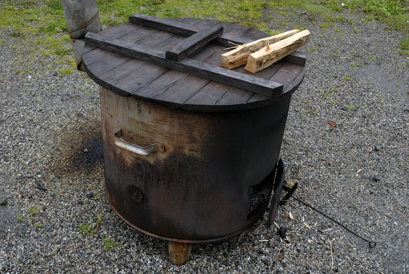 Fût cylindrique en tôle noircie par le feu, poignée rivetée, conduit de fumée à gauche, couvercle fait de planches sur lequel sèchent deux bûchettes, sur du gravier.