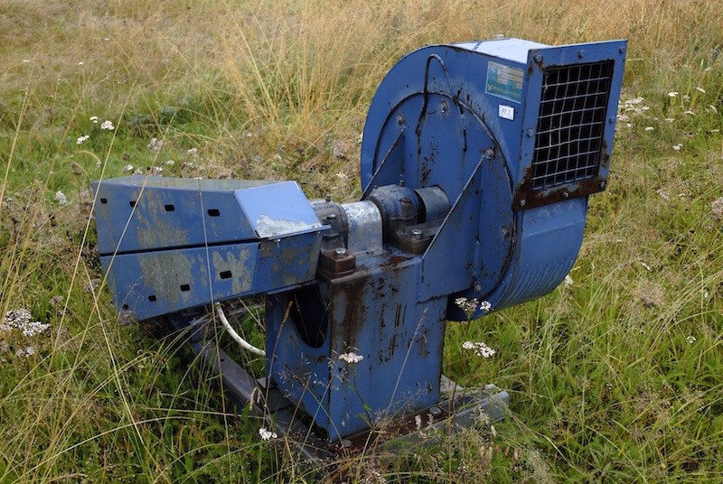 Ventilateur industriel bleu à volute, moteur et carter grillagé, couché dans une prairie d'herbes hautes et d'ombellifères.