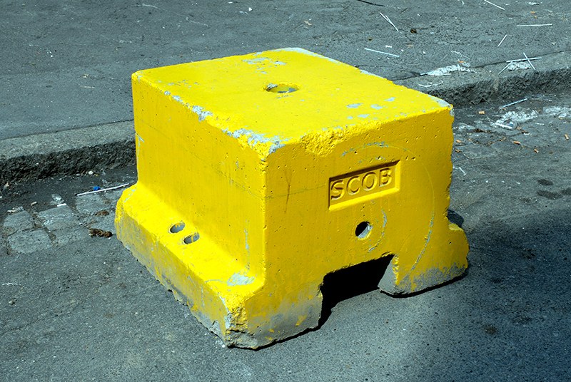 Plot de béton peint en jaune vif, moulé d'un cartouche « SCOB » et percé de trous de manutention, calé contre la bordure d'un trottoir.