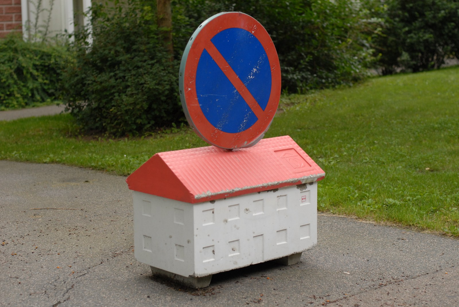 Socle de panneau routier en béton moulé en forme de maison&nbsp;: corps blanc à fenêtres en relief, toit rouge à fausses tuiles, surmonté d'un disque bleu barré de rouge — stationnement interdit. Vue de trois quarts sur une allée.