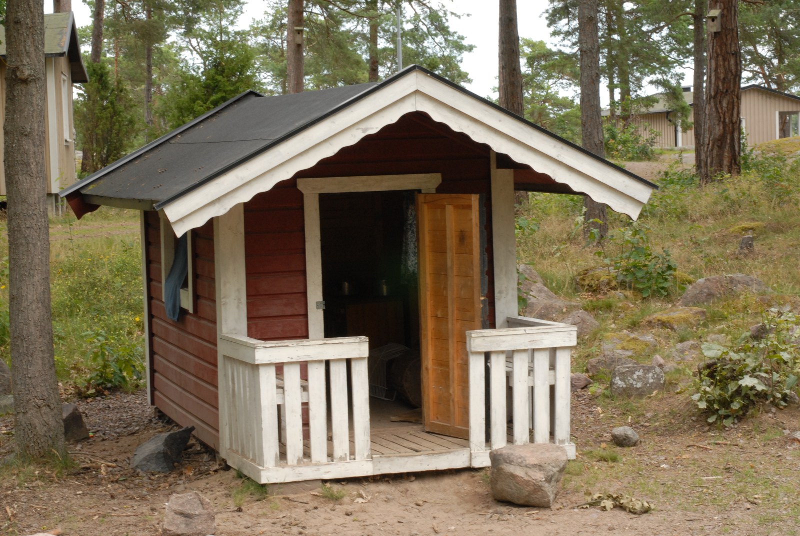 Cabane de jeu en bois rouge sombre à toit noir, avancée couverte, balustrade blanche et porte de bois clair entrouverte, entre les troncs d'une pinède.
