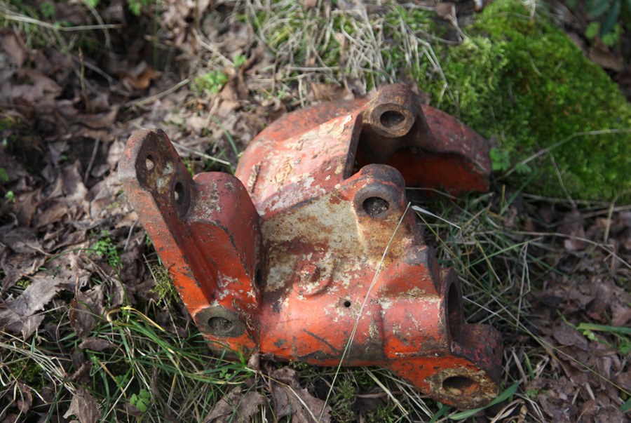 Pièce de fonte orange, épaisse et percée de plusieurs alésages, écaillée et rouillée, posée à même les feuilles mortes et la mousse d'un sous-bois.