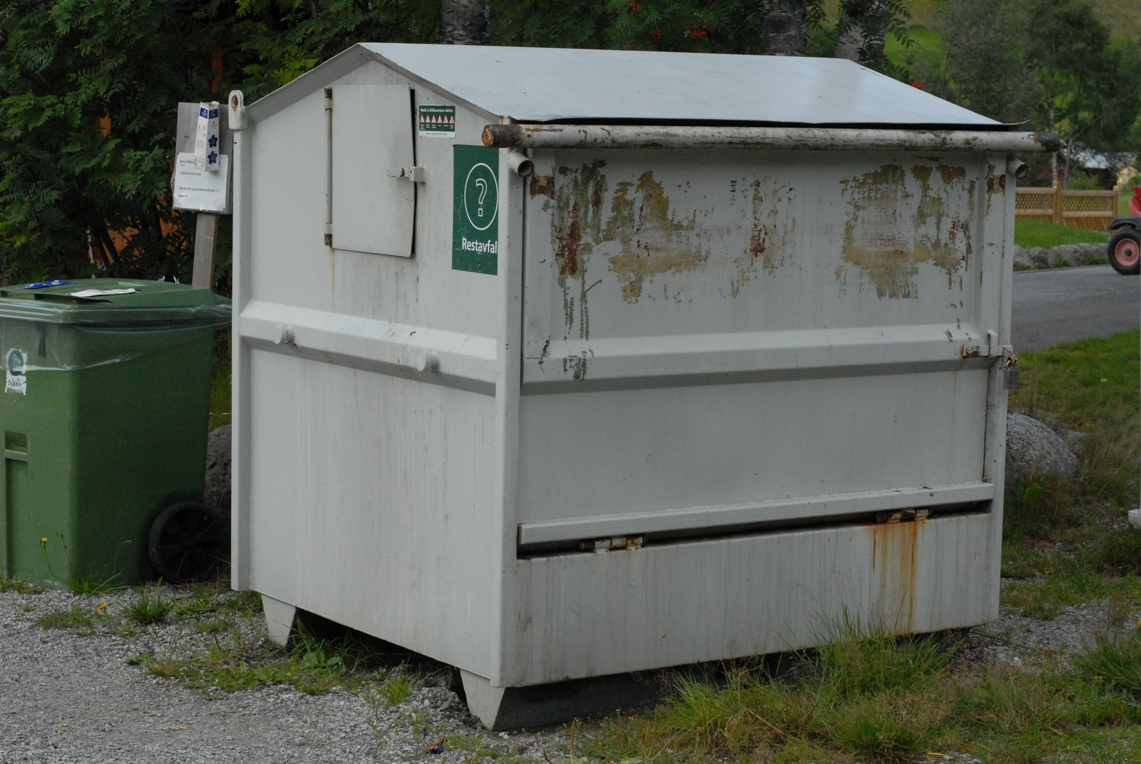 Conteneur à déchets en tôle claire, toit à deux pentes et trappe latérale, une étiquette verte marquée d'un point d'interrogation, à côté d'un bac roulant vert, au bord d'un chemin de gravier.