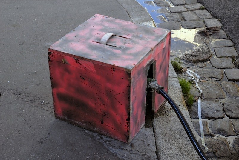 Caisson métallique cubique peint au rouge à la bombe, poignée sur le dessus, un gros câble noir sortant d'une ouverture latérale, posé au bord d'un caniveau pavé.