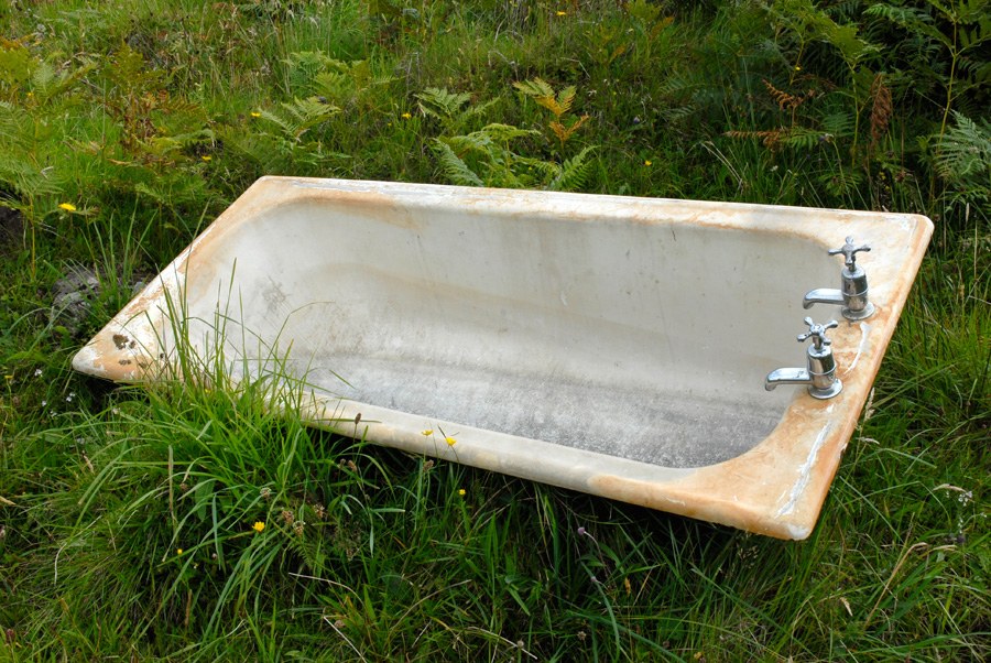Baignoire en fonte émaillée, robinetterie chromée intacte, abandonnée dans une prairie de hautes herbes et de fougères.