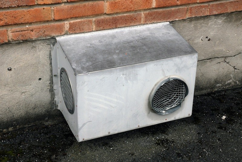Caisson de ventilation en tôle claire, deux ouïes rondes grillagées sur ses flancs et couvercle plat en pente, posé au pied d'un mur de brique.