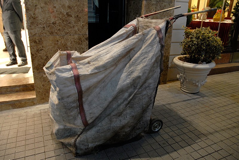 Chariot de nettoyage&nbsp;: un grand sac de toile blanche sanglé de rouge, tendu sur un châssis à deux roues, appuyé contre la façade de marbre d'un immeuble.