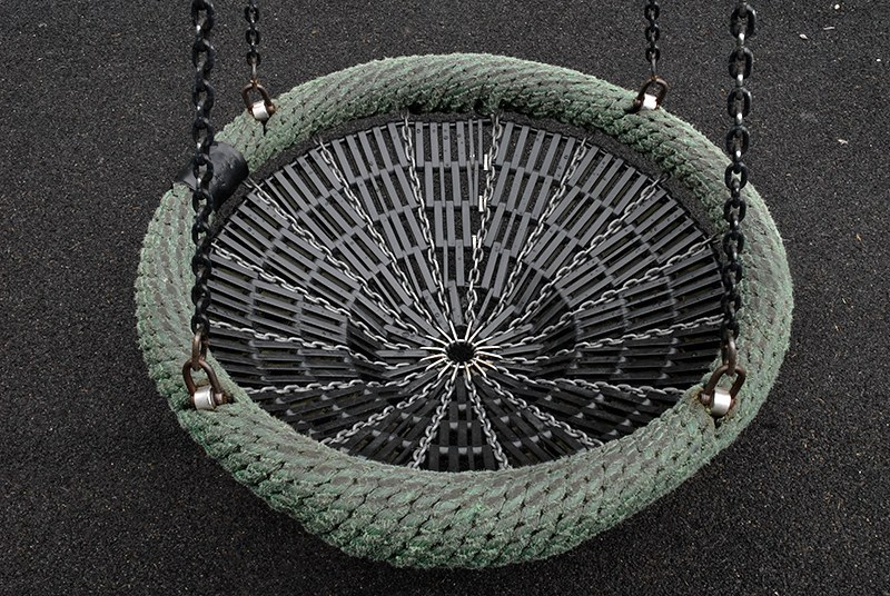 Balançoire-nid vue du dessus&nbsp;: un anneau de cordage vert tressé, tendu d'un tamis de lanières noires rayonnantes, suspendu par quatre chaînes au-dessus d'un sol souple.