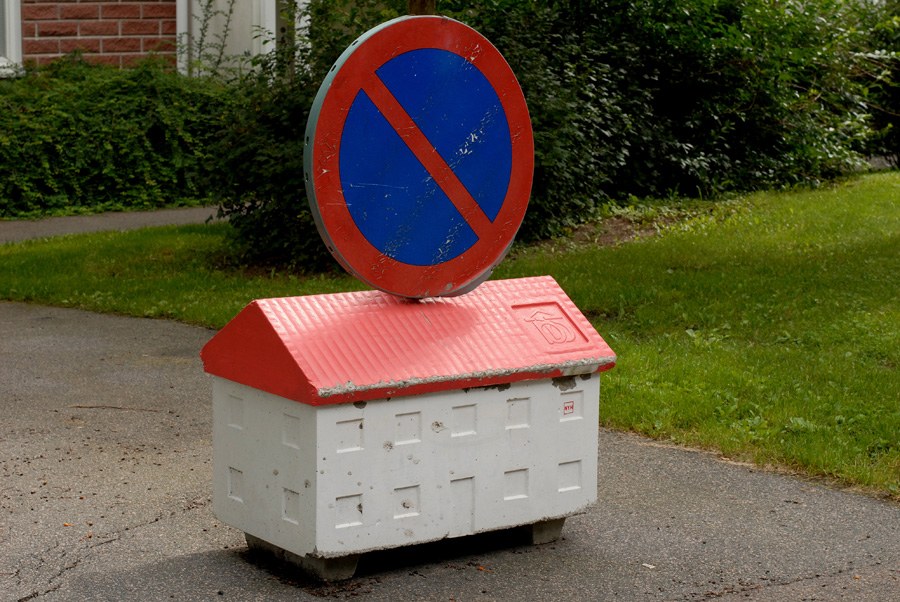 Le même socle-maison de panneau routier, vu de face&nbsp;: corps blanc, toit rouge, surmonté du disque bleu barré de rouge du stationnement interdit, sur une allée bordée de haies.