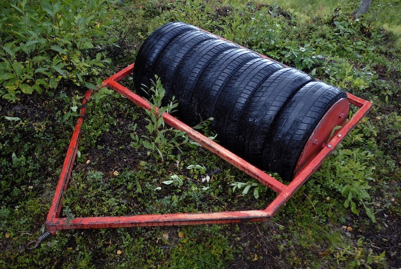 Rouleau de jardin bricolé&nbsp;: une pile de pneus enfilés sur un axe, tenue dans un cadre tubulaire peint en rouge, abandonnée sur un tapis de myrtilliers.