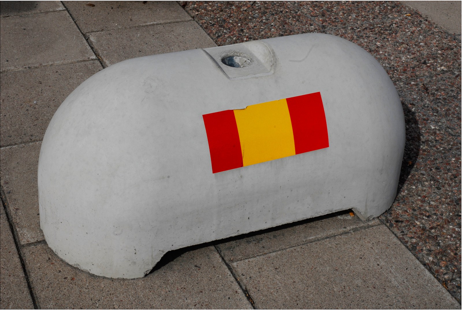 Borne de béton blanche à dos arrondi, marquée d'une bande adhésive rouge-jaune-rouge et d'un œilleton de levage, posée sur un trottoir dallé.