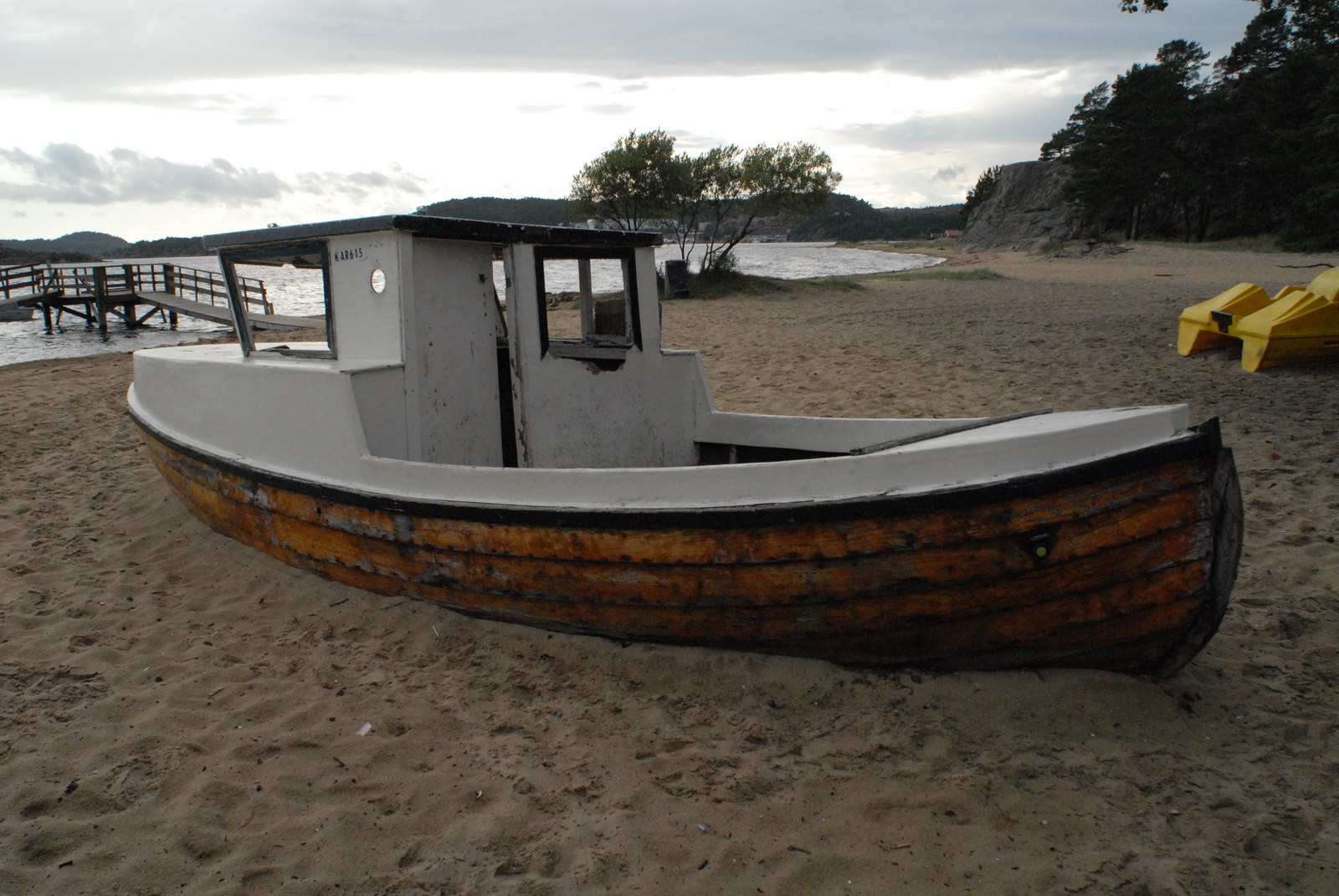 Barque de pêche à cabine, coque orange et noire et superstructure blanche, échouée sur le sable d'une plage, un ponton de bois et des pédalos jaunes à l'arrière-plan.