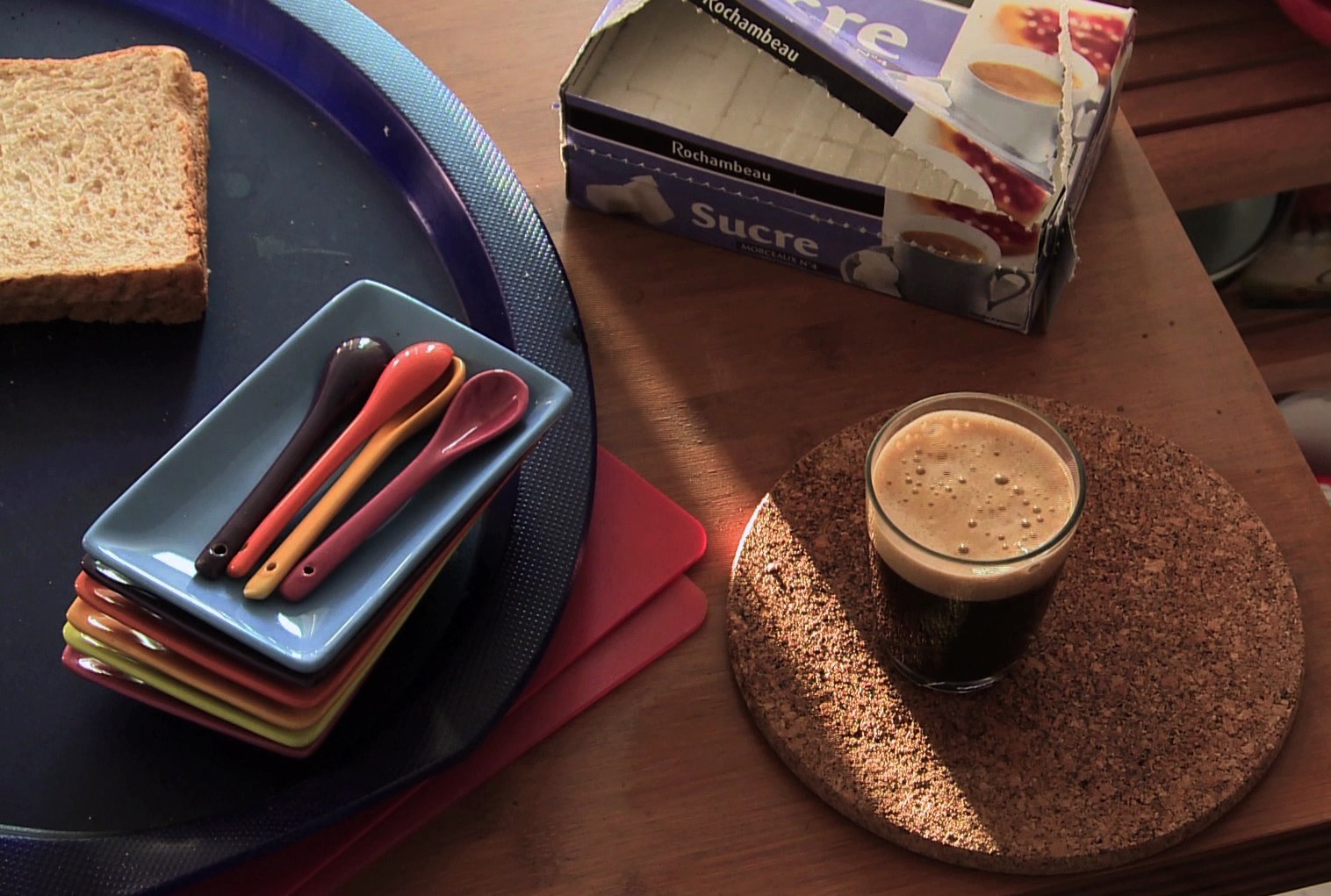 Photogramme : sur une table de bois, un plateau bleu portant du pain de mie et des cuillères de couleur, une boîte de sucre entamée, et un verre de café mousseux posé sur un dessous-de-verre en liège.